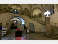 Historischer Innenraum mit einer großen, steinernen Treppe, die mit kunstvollen Ornamenten verziert ist, und mehreren Rundbögen. Im Vordergrund steht eine moderne Skulptur auf einem schwarzen Sockel, der mit einem Text bedruckt ist, und daneben befinden sich Infoständer; die Umgebung ist von warmem Licht beleuchtet.