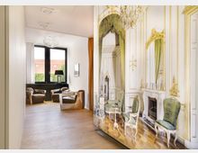 Ein moderner Wohnraum mit hellen Holzböden, großen Fenstern und klassischen Ledersesseln. Im Vordergrund ist eine dekorative Trennwand mit einem aufgedruckten Motiv eines klassisch eingerichteten Salons, einschließlich grüner Polsterstühle, geschnitzter Möbel und Kronleuchter.