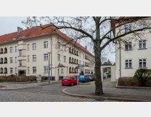 Mehrstöckiges Wohngebäude im Stil des frühen 20. Jahrhunderts mit hellem Putz und rotem Ziegeldach, flankiert von Rundbogenbalkonen. Im Vordergrund eine Baumreihe, parkende Autos und eine kleine Straße mit gepflastertem Gehsteig.