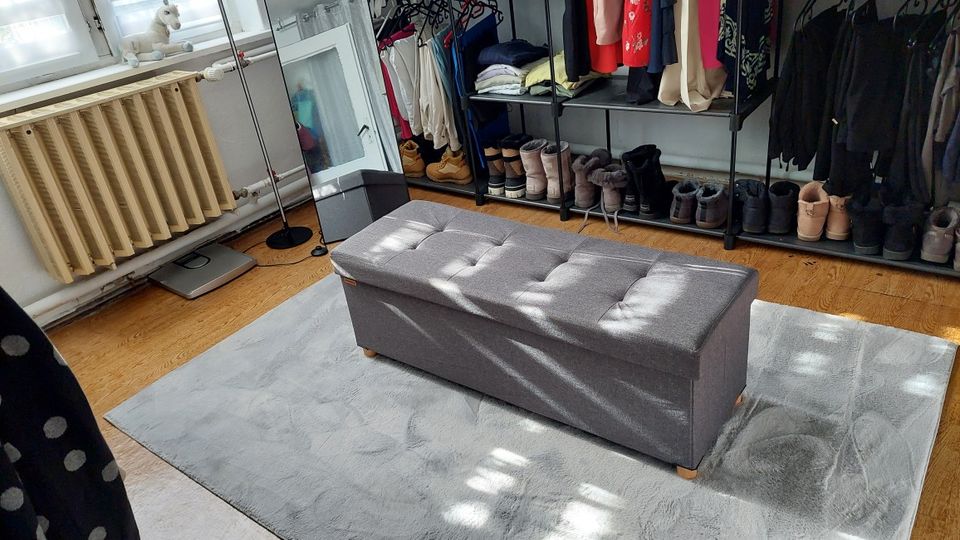 Helles Ankleide- bzw. Garderobenzimmer mit offenem Kleiderregal und Schuhablagen entlang der Wand sowie großen Fenstern über einem Heizkörper. In der Mitte steht eine graue, gepolsterte Sitzbank auf einem hellgrauen Teppich auf Holzboden.