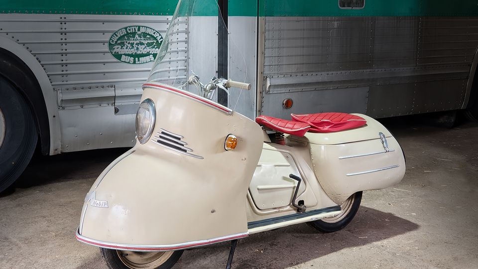 Beiger, stromlinienförmiger Motorroller mit hoher Windschutzscheibe und rotem Doppelsitz steht auf einem Betonboden. Im Hintergrund befindet sich ein silberner, genieteter Bus mit grünem Streifen, was auf eine Garage oder ein Museum mit historischen Fahrzeugen hinweist.