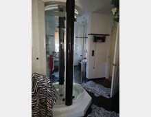 Modernes Badezimmer mit einer zentralen, runden Duschkabine mit schwarzen Säulenelementen und Glastüren. Der Boden ist mit schwarzen Fliesen ausgelegt, teilweise bedeckt von Zebra-Muster-Teppichen; die Wände sind weiß gefliest, ergänzt durch dunkle Zierleisten.