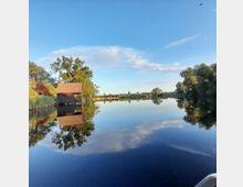 Ruhiger See mit klarer Wasseroberfläche, die den blauen Himmel und umgebende Bäume spiegelt. Links im Bild steht ein Holzhaus auf Pfählen, umgeben von Schilf und Bäumen.