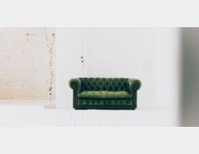 Grünes, gepolstertes Chesterfield-Sofa in einem minimalistischen Raum mit heller, weißer Holzfußbodenoptik. Die Wände bestehen aus einer gemischten weißen und unverputzten Backsteinstruktur.