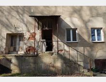 Eingangsbereich eines stark verwitterten Gebäudes mit bröckelndem Putz und freiliegendem Ziegelmauerwerk, einer offenen Tür unter einem kleinen Wellblechvordach. Eine betonierte Rampe mit rostigem Geländer führt zur Tür; daneben sind alte Fenster und überwucherte Bereiche am Sockel zu sehen.