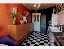 Küche mit schwarz-weiß kariertem Boden, einer weißen Spüle und Arbeitsplatte auf der rechten Seite, einem schwarzen Kühlschrank vor einer weißen Tür und einer roten Wand mit Holzkommode und Dekorationen links. Eine lila Sitzgelegenheit ist teilweise im Vordergrund sichtbar.