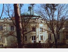 Historische Villa mit cremefarbener Fassade, großen Fenstern und einer grünen Kuppel, umgeben von hohen, kahlen Bäumen. Die Architektur weist klassische Verzierungen und Balustraden auf.