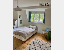 Ein helles Kinderzimmer mit einem Einzelbett, das an einer weißen Wand unter einem großen Fenster mit türkisfarbenen Vorhängen steht. Der Raum hat einen Holzboden, zwei Teppiche (einer rund und geflochten, einer mit Rautenmuster) sowie eine Hängelampe an der Decke.