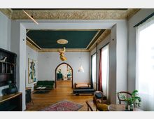 Wohnzimmer mit hohen Decken und einem dunkelgrünen, verzierten Deckenfeld, goldenen Stuckelementen und weißen Wänden. Der Raum enthält eine Mischung aus modernen und klassischen Möbeln, einen Bogendurchgang, große Fenster mit Vorhängen und Holzboden.