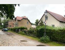 Eine gepflasterte Straße mit Pfützen führt an zwei Wohnhäusern mit roten Ziegeldächern entlang, die von Gärten und Zäunen umgeben sind. Im Vordergrund steht eine historische Straßenlaterne, und am linken Bildrand parken Autos unter Bäumen.