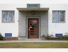 Eingangsbereich eines Wohngebäudes mit einer roten Tür, flankiert von rechteckigen Glasfenstern mit blauen Rahmen. Die Fassade ist weiß mit grauen Beton- und Klinkerelementen, und vor dem Eingang befindet sich ein gepflasterter Weg.