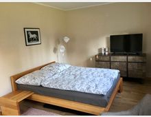 Schlafzimmer mit einem Holzbett, das mit weiß-gemusterter Bettwäsche ausgestattet ist. Im Raum befinden sich eine wandmontierte Zeichnung eines Pferdes, eine Stehlampe, eine Holzanrichte mit Fernseher darauf und ein hellbeigefarbener Anstrich an den Wänden.