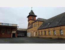 Backsteingebäudekomplex mit langem, ziegelgedecktem Satteldach und Rundbogenfenstern, dahinter ein hoher Turm mit Uhr und spitzem Dach. Davor liegt ein leerer, asphaltierter Hof; an einigen Wänden sind verwitterte Flächen und Graffiti zu sehen.