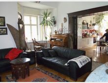 Wohnzimmer mit dunklen Ledersofas, Holzmöbeln und exotischer Dekoration, wie einer großen Skulptur und Wandmasken. Im Hintergrund eine offene Küche mit hellem Tageslicht und modernen Geräten.