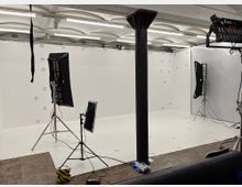 Ein professionelles Fotostudio mit weißen Wänden, markiert mit nummerierten Punkten, ausgestattet mit Beleuchtungseinrichtungen und Stativen. Im Vordergrund befindet sich eine schwarze Säule, während die Decke aus Beton mit schalenförmigen Strukturen gestaltet ist.