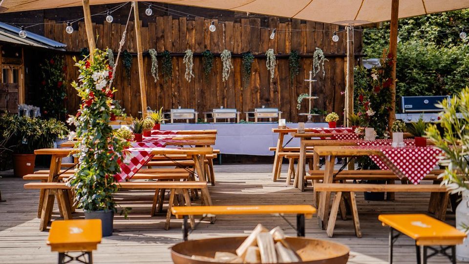 Überdachte Außenterrasse mit Holzboden, langen Bierzeltgarnituren und rot-weiß karierten Tischdecken; darüber hängen Lichterketten und Pflanzenranken. Im Hintergrund steht eine rustikale Holzwand mit Buffetaufbau, seitlich sind Topfpflanzen und ein Grill zu sehen.