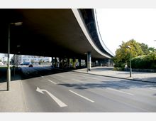 Straßenunterführung mit einer breiten, geschwungenen Brücke, die von Betonpfeilern gestützt wird. Im Vordergrund verläuft eine mehrspurige Straße, daneben ein Fußweg mit Laternen und Bäumen im Hintergrund.