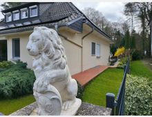 Einfamilienhaus mit heller Fassade und dunklem Dach, im Vordergrund eine weiße Löwenstatue aus Stein auf einem Pfeiler. Der Garten ist gepflegt mit grünem Rasen, Sträuchern und einem Metallzaun, im Hintergrund sind Bäume sichtbar.