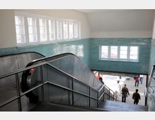 Ansicht eines U-Bahn-Eingangsbereichs mit einer Rolltreppe und hellblauen Fliesen an den Wänden. Durch Fenster dringt Tageslicht, und im unteren Bereich sind Fußgänger sowie ein Hinweisschild zu den Stationen "Nollendorfplatz" und "Krumme Lanke" zu sehen.