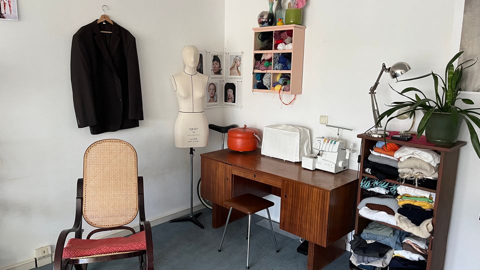 Small sewing and craft studio corner with a wooden desk holding a serger and covered machine, a dress form beside it, and wall-mounted cubbies filled with yarn. A rocking chair, a hanging jacket, a shelving unit stacked with folded fabric, and a desk lamp with potted plants sit against white walls on a carpeted floor.