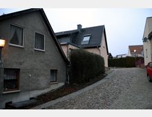 Gepflasterte Straße in einem Wohngebiet mit mehreren Einfamilienhäusern, darunter ein graues und ein beige gestrichenes Haus mit steilen Dächern. Ein Laternenmast und eine Hecke flankieren die Straße, daneben steht ein rotes Auto geparkt.