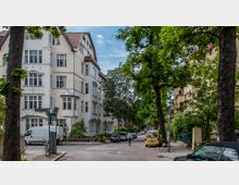 Straßenszene in einem Wohngebiet mit mehrstöckigen, hellen Altbauten im Jugendstil. Die Straße ist von Bäumen gesäumt, parkende Autos und ein Gehweg sind sichtbar; im Hintergrund läuft eine Person auf der leicht geschwungenen Straße.