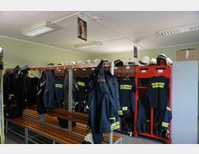 Umkleideraum einer Feuerwehrstation mit Holz-Sitzbänken, offenen Spinden und Kleiderhaken. Feuerwehrkleidung, Helme und schwarze Shirts mit der Aufschrift "Feuerwehr Vierraden" sind sichtbar aufgehängt oder verstaut.