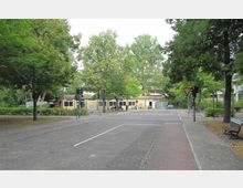 Straßenszene mit Fußgängerüberweg und Ampeln, umgeben von Bäumen. Im Hintergrund steht ein flaches, gelbes Gebäude mit mehreren Fenstern; davor sind Fahrräder abgestellt und eine Sitzgruppe erkennbar.