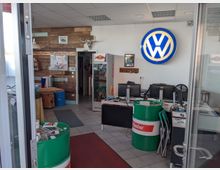 Ein kleines Büro oder Verkaufsraum mit Autowerkstätten-Elementen, darunter ein großes beleuchtetes Volkswagen-Logo an der Wand und ein grünes Castrol-Fass als Dekoration. Holzvertäfelte Wand mit Deko, mehrere Schreibtische mit Computern und eine Glasvitrine mit Produkten im Hintergrund.