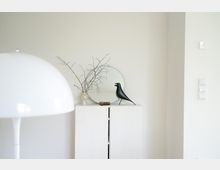 Minimalistisch eingerichteter Innenraum mit hellem Wandschrank, darauf ein runder Spiegel, eine Vase mit kahlen Zweigen und eine schwarze Vogelfigur. Im Vordergrund eine weiße, halbrunde Lampe, rechts an der Wand Lichtschalter.