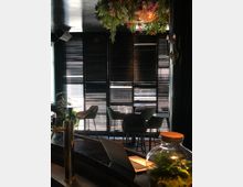 Eine stilvolle Innenraumaufnahme mit dunklen Jalousien, die das Licht filtern, Barhockern und runden Tischen im Hintergrund. Im Vordergrund stehen ein geschwungener Tresen mit einer Zapfsäule, ein aufgeklappter Laptop, und ein Glasgefäß mit Pflanzen; von der Decke hängt ein Kronleuchter mit Blumenarrangement.