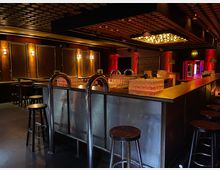 Eine stilvolle Bar mit metallischer Theke, auf der gestapelte Gläser stehen, umgeben von hohen Hockern. Die dunkelrote Beleuchtung, Holzelemente und eine Decke mit runden Ornamenten prägen den Raum, der eine gemütliche, moderne Atmosphäre ausstrahlt.