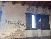 Innenansicht eines historischen Gebäudes mit unverputzten, teils beschädigten Ziegelwänden, auf denen handgeschriebene alte Inschriften sichtbar sind. Zwei große Fenster mit dunklen Holzläden und abgenutzten Rahmen lassen Tageslicht ins Zimmer, während Holzbalken die Decke stützen.