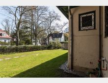 Seitenansicht eines Wohnhauses mit verputzter Fassade und vergittertem Fenster, daneben ein gepflegter Garten mit Rasen, vereinzelten Steinplatten als Weg und einem Holzzaun im Hintergrund. Im Hintergrund sind weitere Häuser und Bäume sichtbar.