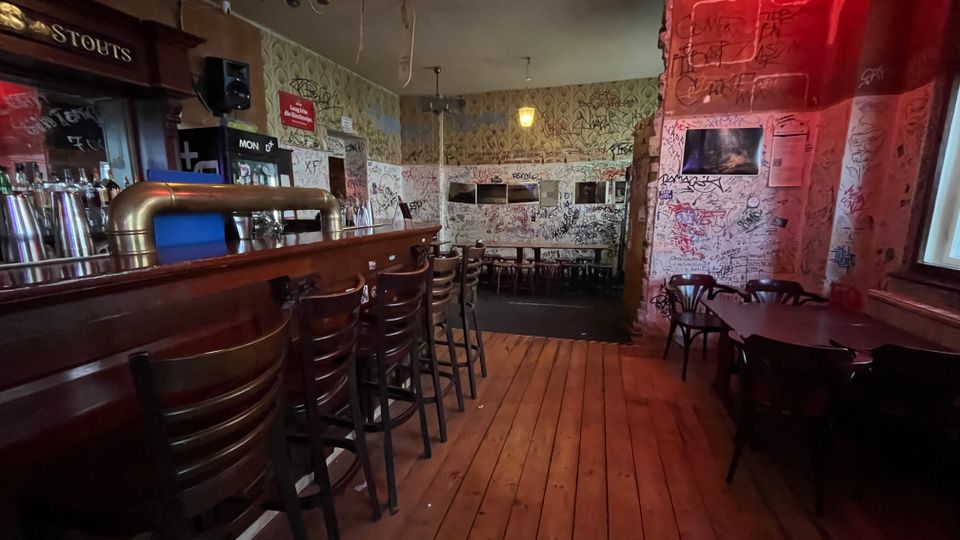 Innenraum einer schummrigen Kneipe mit langer Holztheke und Barhockern links, Holzdielenboden und Tischen mit Stühlen rechts. Die Wände sind dicht mit Graffiti und Kritzeleien bedeckt, beleuchtet von warmen Deckenlampen und rotem Licht im vorderen Bereich.