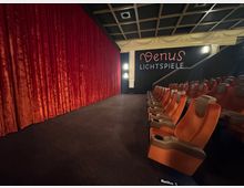 Innenansicht eines Kinosaals mit orangefarbenen Polstersesseln und roten Samtvorhängen vor der Leinwand. Im Hintergrund ist eine Wand mit der neonbeleuchteten Aufschrift „Venus Lichtspiele“ und dekorativen Säulen zu sehen.