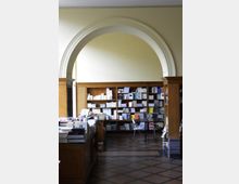 Innenraum einer Buchhandlung mit hohen, hölzernen Regalen, die mit Büchern und Zeitschriften gefüllt sind, sowie einem markanten Rundbogen an der Wand. Im Vordergrund steht ein Holztresen, und der Boden ist mit einem geometrischen Muster gestaltet.