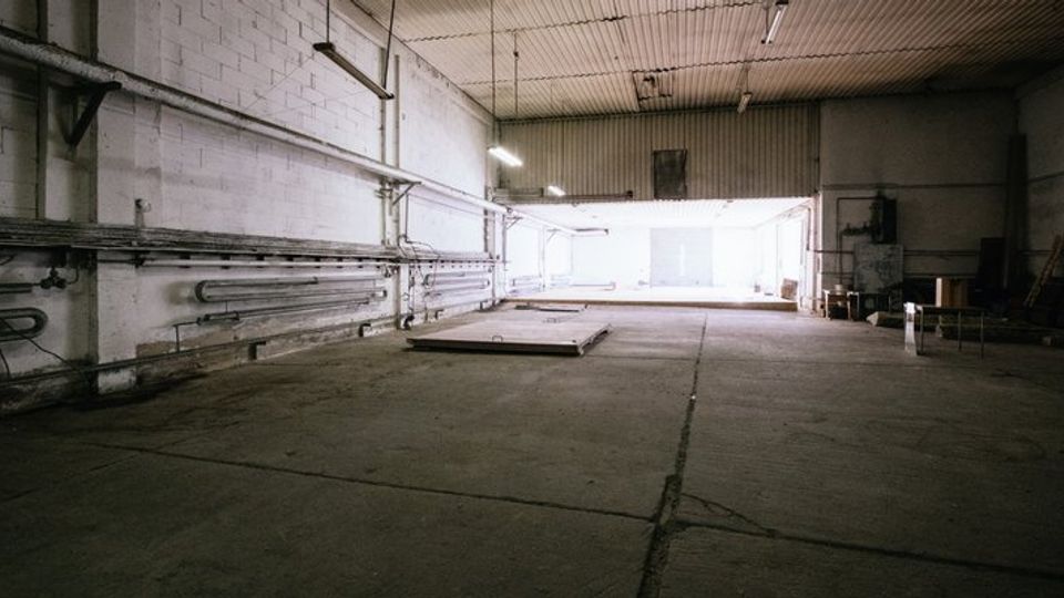 Ein leerer, großer Industrie- oder Lagerraum mit Betonboden, hohen Wänden aus weiß gestrichenen Ziegeln, Deckenbeleuchtung und einigen sichtbaren Rohrleitungen entlang der Wände. Im Hintergrund ist ein großes, von natürlichem Licht erhelltes Tor oder Ausgang zu sehen.