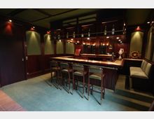 Elegante Bar mit dunkler Holzvertäfelung, einer langen Theke mit vier gepolsterten Barhockern und hinterleuchteten Regalen, die Flaschen und Gläser präsentieren. Die Einrichtung umfasst nautische Dekorelemente wie ein Steuerrad und gedämpfte Beleuchtung für eine warme Atmosphäre.