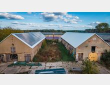 Ehemaliger landwirtschaftlicher Hof mit zwei langgezogenen Gebäuden, die sich gegenüberliegen. Das linke Gebäude hat ein Dach mit Solarpanelen, das rechte zeigt bunte Graffitis an der Außenwand; dazwischen befindet sich ein überwucherter Hof mit Vegetation. Im Hintergrund ist Wald zu sehen.