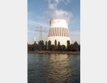 Ein großes Kühlturm eines Kraftwerks mit vertikalen braunen Streifen, umgeben von einer Reihe von Bäumen. Im Vordergrund ist ein Fluss sichtbar, und links vom Kühlturm steht ein Strommast.