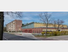 Ein Wohngebäude mit vier Etagen aus rotem Backstein steht links, daneben eine flache Sporthalle mit Wellblechdach und gelben Wänden. Im Vordergrund sind ein Gehweg, eine Straße, ein roter Bushaltestellenunterstand und einige Bäume sichtbar.