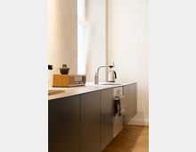 Moderne Küche mit schlichtem Design, bestehend aus einer dunklen Unterschrankzeile, einer hellen Arbeitsplatte, einem eingebauten Ofen und einem Wasserhahn mit Kessel. Im Hintergrund befindet sich ein Fenster, das Tageslicht in den Raum einlässt.