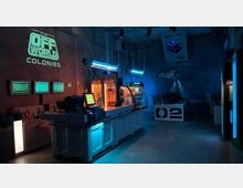 Futuristisch gestalteter Innenraum mit blauem Licht beleuchtet, featuring eine Snack-Theke mit leuchtenden Kühlschränken und Displays. Im Hintergrund befinden sich Arbeitsstationen mit Bildschirmen und eine große Lüftungsanlage an der Decke.