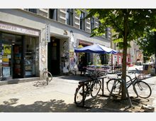 Straßenszene mit einem Buchladen in einem Altbau, daneben ein Café mit blauen Sonnenschirmen und Sitzbänken im Außenbereich. Fahrräder stehen an einem Fahrradständer, umgeben von Bäumen und Pflastersteinen.