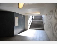 Unterführung mit gefliesten Wänden aus hellen und dunklen Kacheln, Deckenleuchten und einer Treppe, die nach oben ins Freie führt. Auf der linken Wand ist ein leerer Schaukasten angebracht.