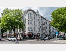 Eckgebäude im Gründerzeitstil mit weißen Fassaden und dekorativen Verzierungen, im Erdgeschoss befinden sich Geschäfte und ein Café mit Außensitzplätzen. Umgeben von großen Bäumen und einer belebten Straßenkreuzung mit Fußgängern, Fahrradfahrern und geparkten Autos.