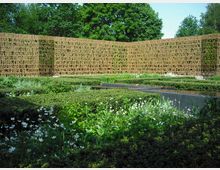Gartenanlage mit niedrigen Hecken und Beeten voller weißer Blüten, in der Mitte ein rechteckiges Wasserbecken mit dunkler Einfassung. Dahinter steht eine lange, gebogene Sichtwand aus goldfarbenem Metallgitter mit ausgeschnittenen Buchstaben, umgeben von hohen Bäumen.