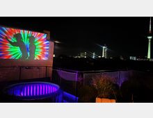 Rooftop-Terrasse bei Nacht mit Blick auf den Berliner Fernsehturm und Stadtlichter. Im Vordergrund eine beleuchtete aufblasbare Wanne, eine Netzabgrenzung sowie eine Wandprojektion mit bunten Lichtstrahlen und Silhouetten von tanzenden Personen.