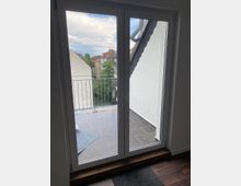 Blick durch eine Glastür auf einen kleinen Balkon mit dunklen Fliesen, einem Metallgeländer und einer seitlichen Hauswand. Im Hintergrund sind Nachbargebäude, Bäume und ein teilweise bewölkter Himmel sichtbar.